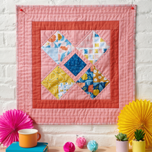 Load image into Gallery viewer, Colorful quilt on a white brick wall with decorative items below