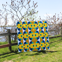 Load image into Gallery viewer, Colorful geometric-patterned quilt on a grassy field with a wooden fence and trees in the background.